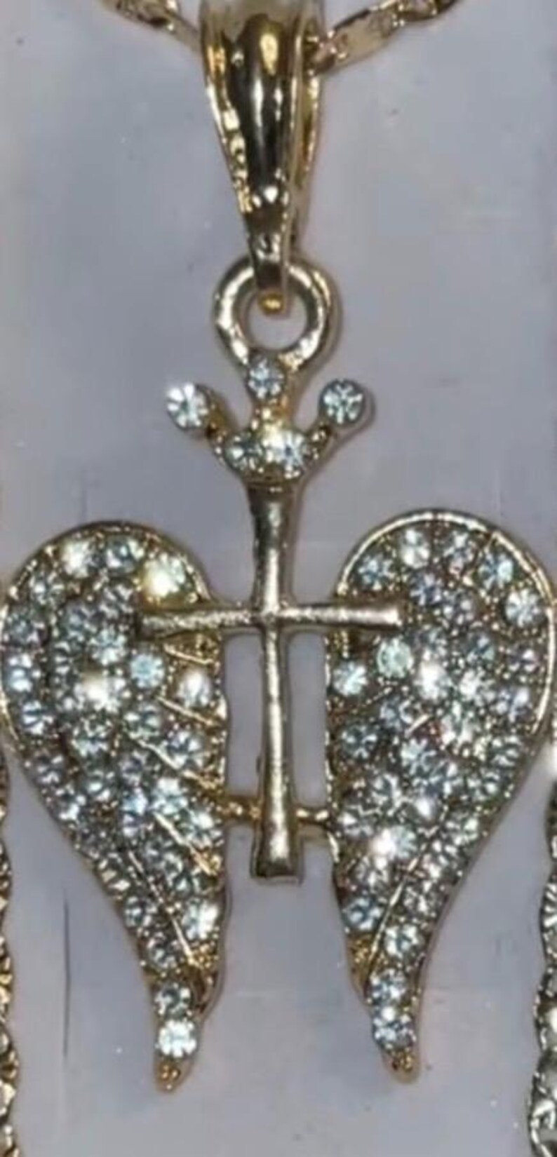 May include: Gold-coloured pendant necklace with a cross design and two heart-shaped wings, both set with sparkling crystals. The pendant has a decorative bail and is likely made of a metal alloy.