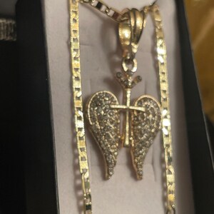 May include: Gold-coloured necklace with a pendant featuring a heart-shaped design with wings and a cross. The pendant is embellished with small, sparkling stones. The necklace is displayed in a black box with a light-coloured interior.