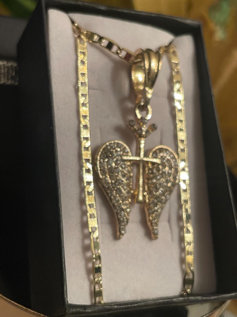 May include: Gold-coloured necklace with a pendant featuring a heart-shaped design with wings and a cross. The pendant is embellished with small, sparkling stones. The necklace is displayed in a black box with a light-coloured interior.