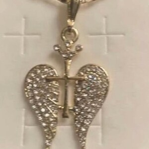 May include: Gold-coloured pendant necklace with a cross design and angel wings, set with small, sparkling stones. The necklace has a fine chain and is shown against a white backdrop.