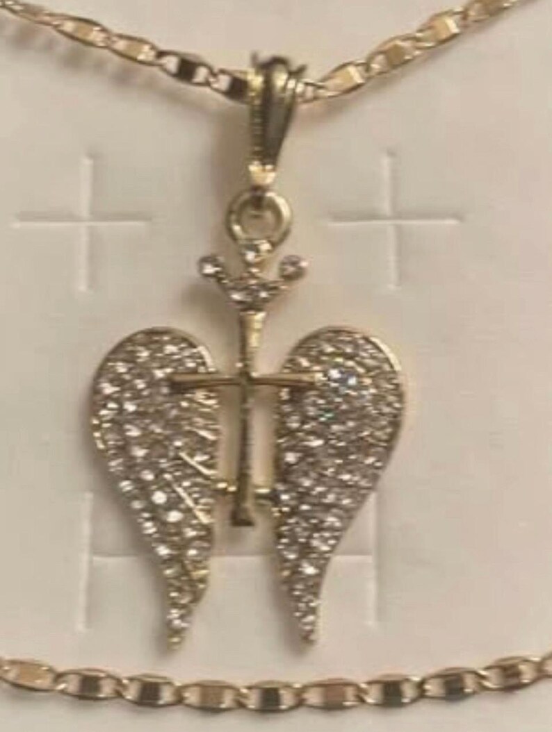 May include: Gold-coloured pendant necklace with a cross design and angel wings, set with small, sparkling stones. The necklace has a fine chain and is shown against a white backdrop.