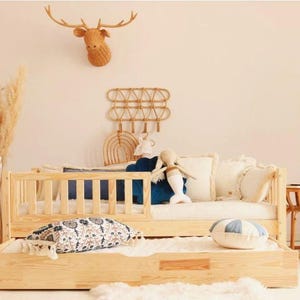 Peut inclure: Lit pour tout-petits en bois clair avec un lit gigogne, avec un matelas blanc, des coussins bleus et des oreillers décoratifs. Une poupée sirène et un coussin rond bleu et blanc sont sur le lit. Une tête de cerf décorative est accrochée au mur.