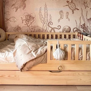 Op de afbeelding: Een licht houten peuterbed met een uitschuifbare lade. Het bed heeft een lattenbodem en voeteneinde. Een schelpvormige mobiel hangt aan de zijkant. Het beddengoed omvat een dekbed met patroon en een halve maan kussen. De muur heeft een zeedieren muurschildering.