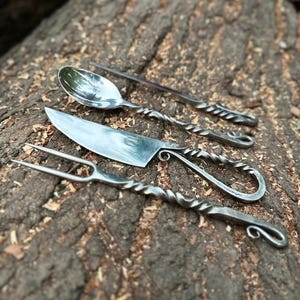 May include: A set of four handcrafted metal utensils: a spoon, a knife, a fork, and a serving spoon. The handles have a twisted design, and the utensils are displayed on a textured, brown wooden surface.