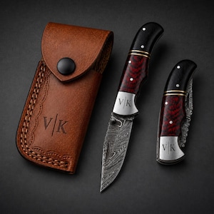 May include: A brown leather sheath with a snap closure and the letters V|K embossed on the front, alongside two folding knives. Both knives feature red and black patterned handles, silver bolsters engraved with V|K, and Damascus steel blades.