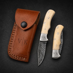 May include: A brown leather sheath with a black snap button and the letters V|K embossed on the front, alongside two folding knives with cream-coloured handles, Damascus steel blades, and the letters V|K engraved on the metal bolsters of each knife.
