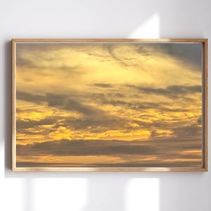 May include: A rectangular framed wall art piece featuring a photograph of a sunset sky. The image displays layers of golden yellow and orange clouds interspersed with dark grey cloud formations, all enclosed within a light-toned wooden frame.