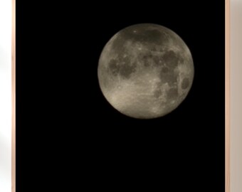 May include: A framed print featuring a full moon against a black background. The moon is a pale, textured sphere, with visible craters and shadows. The frame is a light wood color.