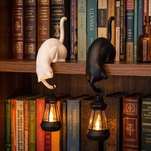 May include: Two cat-shaped ornaments, one white and one black, perched on a shelf edge. Each cat has a curved tail holding a small, black, battery-operated lantern with a warm, glowing light inside, positioned in front of a bookshelf.
