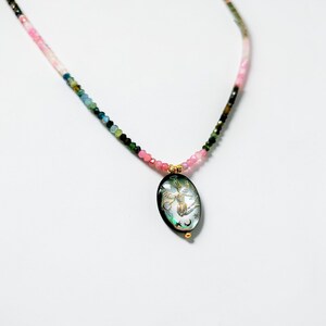 May include: A beaded necklace with a pendant. The necklace features a gradient of pink, green, and black beads. The oval pendant has a black background with a gold design of a bird, moon, and stars. The pendant is accented with gold details.