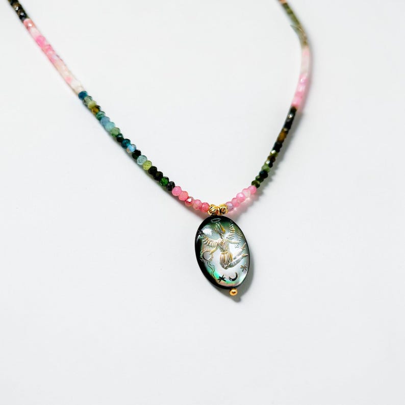 May include: A beaded necklace with a pendant. The necklace features a gradient of pink, green, and black beads. The oval pendant has a black background with a gold design of a bird, moon, and stars. The pendant is accented with gold details.