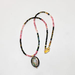May include: A beaded necklace with a gold clasp and a pendant. The necklace features a variety of small, colorful beads in shades of pink, green, black, and yellow. The pendant is oval-shaped with a dark, iridescent surface.