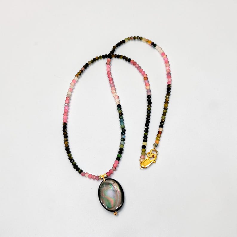 May include: A beaded necklace with a gold clasp and a pendant. The necklace features a variety of small, colorful beads in shades of pink, green, black, and yellow. The pendant is oval-shaped with a dark, iridescent surface.