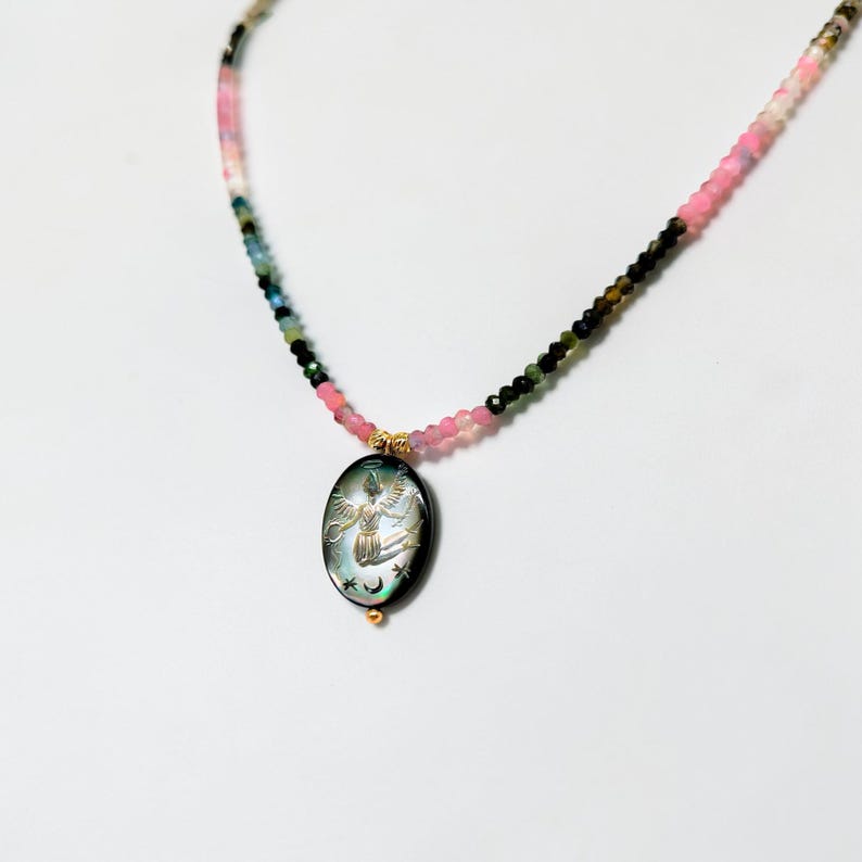 May include: A beaded necklace with a pendant. The necklace features a mix of pink, green, and black beads. The pendant is an oval shape with an angel design. The pendant is black with iridescent colors and gold accents.