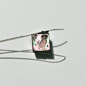 May include: A small, iridescent square with an owl illustration held by tweezers. The owl is depicted with detailed feathers and a crescent moon. The square also features Greek letters and an olive branch design.