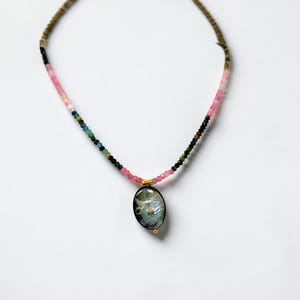 May include: A beaded necklace with a pendant. The necklace features a gradient of pink, green, and black beads. The pendant is an oval shape with a silver design and gold accents. The necklace is on a white background.