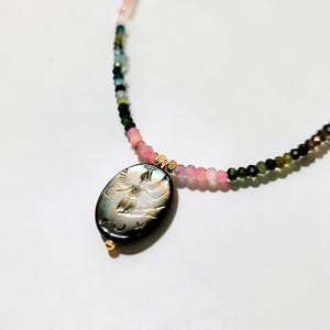 May include: A beaded necklace featuring a pendant with an engraved angel design. The necklace has a mix of pink, green, and black beads. The pendant is oval-shaped with gold accents.