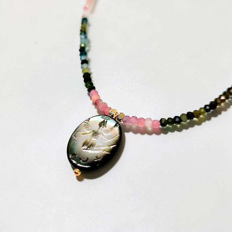 May include: A beaded necklace featuring a pendant with an engraved angel design. The necklace has a mix of pink, green, and black beads. The pendant is oval-shaped with gold accents.