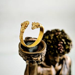 Może przedstawiać: Otwarta bransoletka w kolorze złotym z detalami w kształcie głowy barana na każdym końcu. Bransoletka jest prezentowana na ozdobnym stojaku z ciemnym kamieniem w środku. Tło jest rozmyte.