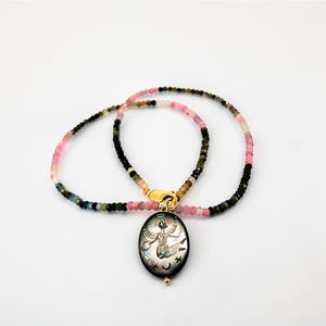 May include: A beaded necklace with a pendant. The necklace features a mix of pink, green, and black beads. The pendant is oval-shaped, black, and has an engraved angel design with stars and moons. The clasp is gold-colored.