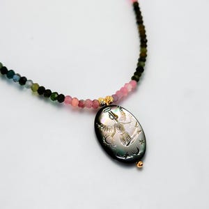 May include: A necklace featuring a pendant with an engraved angel design. The pendant is black with a pearlescent sheen, and the necklace has a beaded strand in shades of pink, green, and gold. The necklace is a delicate piece of jewelry.