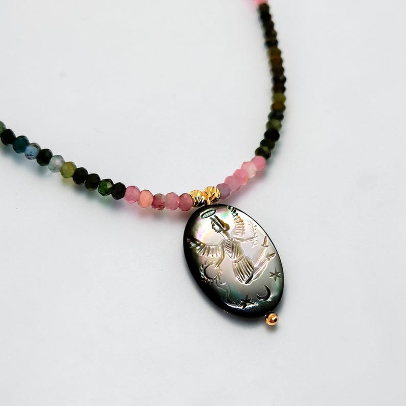 May include: A necklace featuring a pendant with an engraved angel design. The pendant is black with a pearlescent sheen, and the necklace has a beaded strand in shades of pink, green, and gold. The necklace is a delicate piece of jewelry.