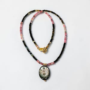 May include: A beaded necklace with a gold clasp and a pendant. The necklace features a mix of pink, green, and black beads. The oval pendant has a carved design of a figure with outstretched arms.