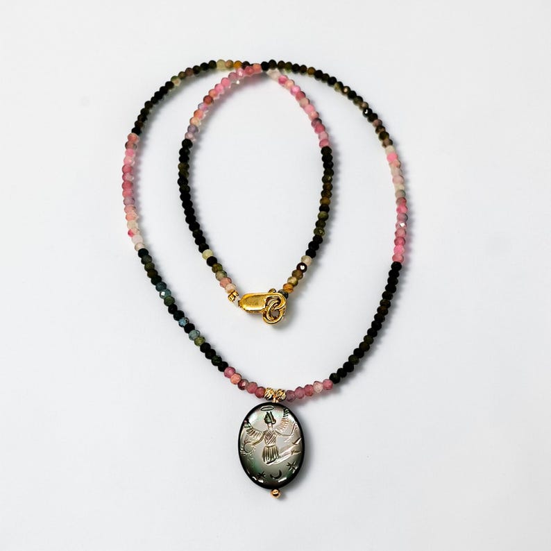 May include: A beaded necklace with a gold clasp and a pendant. The necklace features a mix of pink, green, and black beads. The oval pendant has a carved design of a figure with outstretched arms.