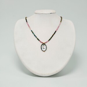 May include: A delicate necklace featuring a pendant with a detailed design. The necklace has a strand of small, colorful beads in shades of pink, green, and blue. The pendant is oval-shaped with a gold-colored frame.