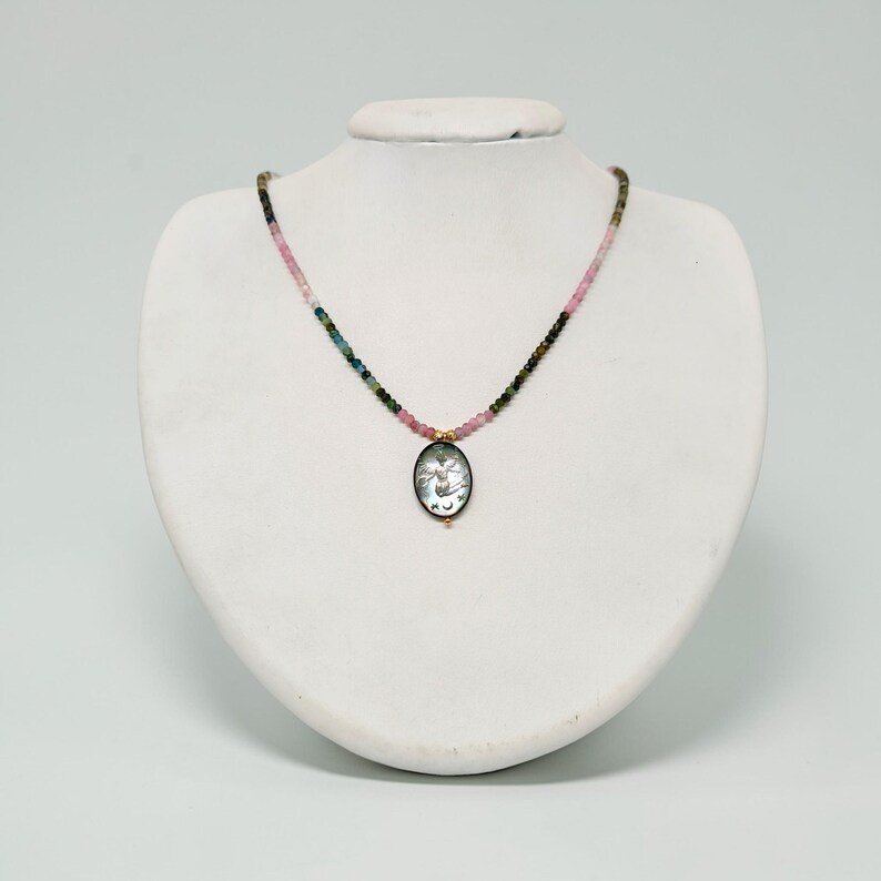 May include: A delicate necklace featuring a pendant with a detailed design. The necklace has a strand of small, colorful beads in shades of pink, green, and blue. The pendant is oval-shaped with a gold-colored frame.