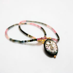 May include: A delicate necklace with a pendant featuring an engraved design. The necklace has a beaded strand in shades of pink, green, and black. The pendant is oval-shaped with a gold-colored bail and a small pink accent.