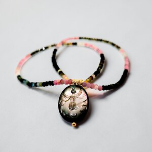 May include: A necklace with a black oval pendant featuring an engraved design of a figure with outstretched wings, stars, and a crescent moon. The necklace has a beaded chain with pink, black, and multicolored beads. The pendant has a gold-colored bail and a small gold bead at the bottom.