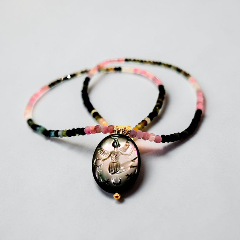 May include: A necklace with a black oval pendant featuring an engraved design of a figure with outstretched wings, stars, and a crescent moon. The necklace has a beaded chain with pink, black, and multicolored beads. The pendant has a gold-colored bail and a small gold bead at the bottom.