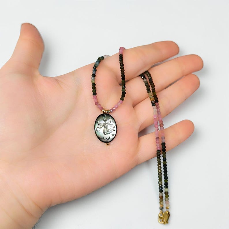 May include: A delicate necklace featuring a pendant with an engraved angel design, accented with stars and moons. The necklace has a beaded chain with pink, green, and black beads. The pendant is oval-shaped with gold-colored accents.