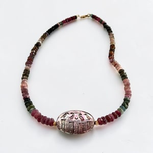 Op de afbeelding: Een ketting met een gouden sluiting. De ketting heeft een verscheidenheid aan kleurrijke edelstenen, waaronder roze, groene en zwarte kralen. Het middelpunt is een ovale hanger met een gegraveerd ontwerp van vogels en architectonische elementen.