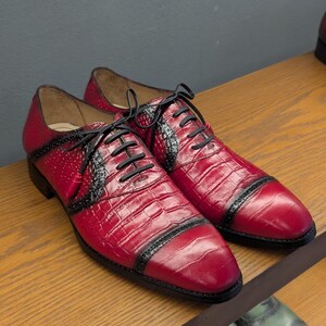May include: A pair of red and black oxford shoes featuring a textured crocodile-skin pattern, black laces, and decorative brogue detailing along the toe cap and side panels. The shoes have a pointed toe design and a low heel with a polished finish.