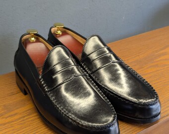 May include: A pair of black leather penny loafers with a glossy finish. The shoes have a classic design with a stitched vamp and a leather sole. The interior is lined with red leather. Shoe trees are inserted in the shoes.