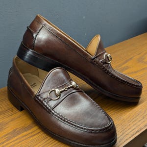 May include: A pair of brown leather loafers with a classic design. Each shoe features a decorative metal bit across the vamp. The shoes are displayed on a wooden surface, showcasing their polished appearance and quality craftsmanship.
