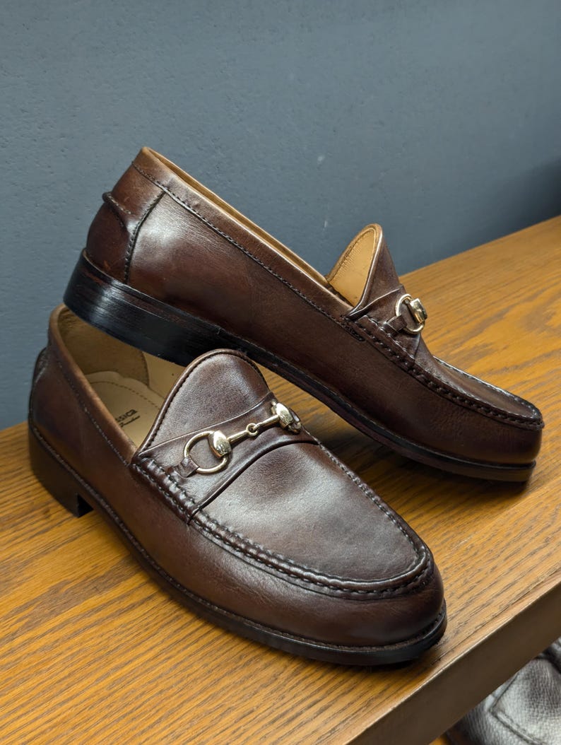 May include: A pair of brown leather loafers with a classic design. Each shoe features a decorative metal bit across the vamp. The shoes are displayed on a wooden surface, showcasing their polished appearance and quality craftsmanship.