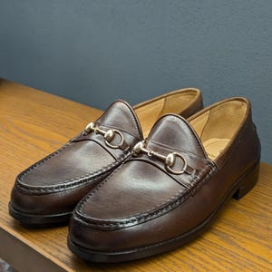 May include: A pair of brown leather loafers with gold-colored horsebit hardware. The shoes are displayed on a wooden surface against a muted blue-gray background. The loafers have a classic design with visible stitching.