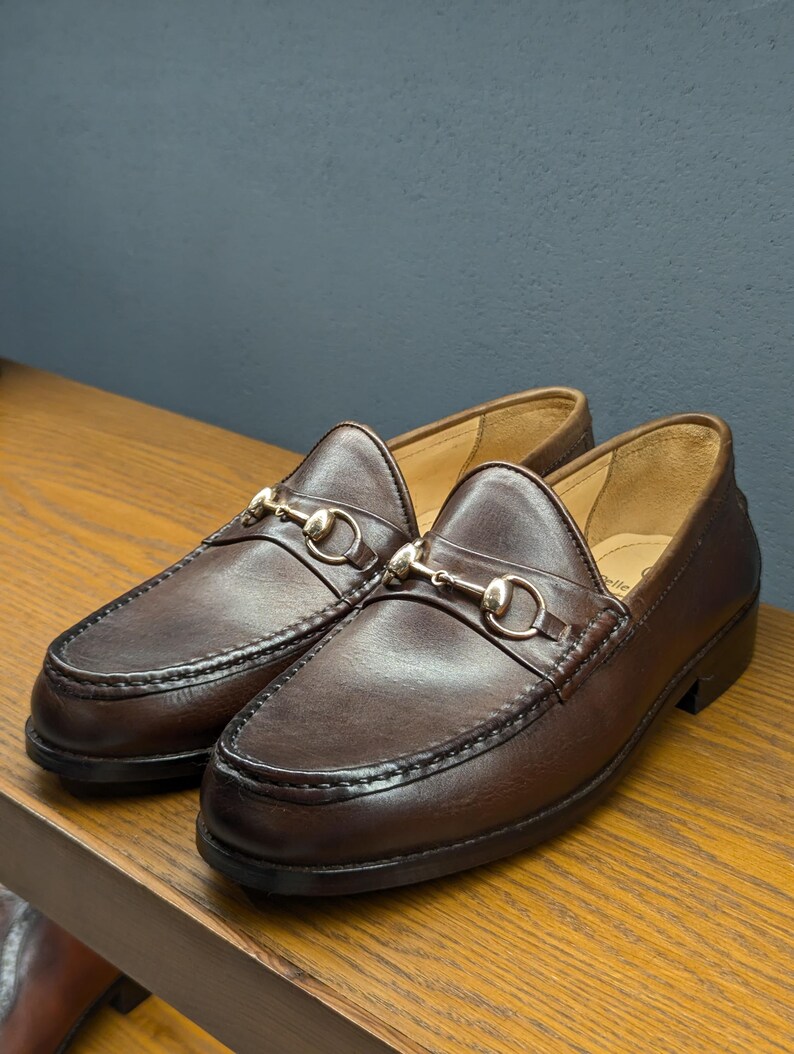 May include: A pair of brown leather loafers with gold-colored horsebit hardware. The shoes are displayed on a wooden surface against a muted blue-gray background. The loafers have a classic design with visible stitching.