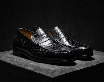 May include: A pair of black leather loafers with a crocodile skin texture. The shoes are displayed on a dark gray surface against a black background. The loafers have a classic design with a rounded toe and a low heel.