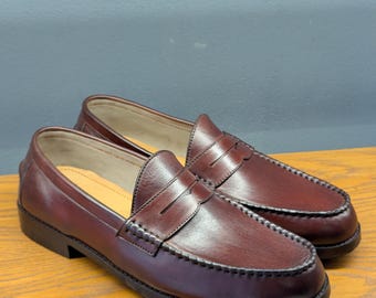 May include: A pair of burgundy leather penny loafers. The shoes have a classic design with a rounded toe, a strap across the vamp, and a stitched welt. The soles are black, and the interior is a light tan color.