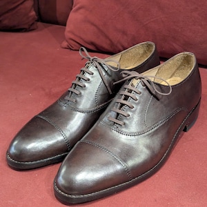May include: A pair of dark brown leather oxford shoes featuring a cap toe design, tonal stitching, and matching dark brown laces. The shoes have a smooth, polished finish with a low heel and a tan interior lining visible at the collar.