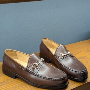 May include: A pair of brown leather loafers with a gold-colored metal horsebit detail. The shoes have a classic slip-on design with a rounded toe and a stitched sole. The loafers are displayed on a wooden surface.