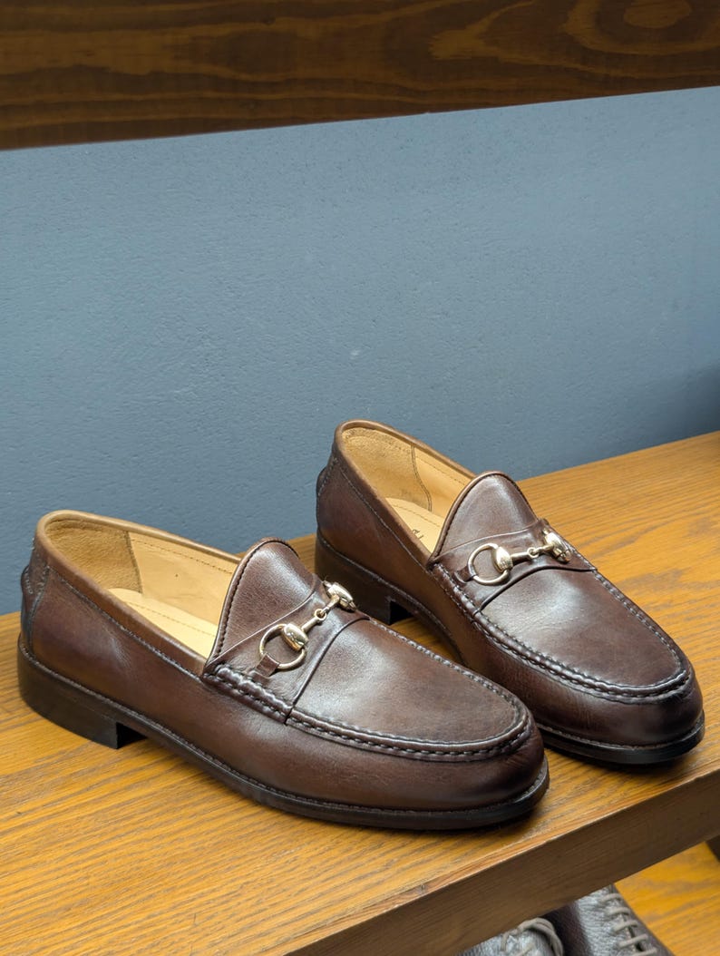 May include: A pair of brown leather loafers with a gold-colored metal horsebit detail. The shoes have a classic slip-on design with a rounded toe and a stitched sole. The loafers are displayed on a wooden surface.