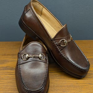 May include: A pair of brown leather loafers with gold-colored metal accents. The shoes are displayed on a wooden surface, showcasing their classic design and stitching details. The loafers feature a slip-on style with a decorative strap.