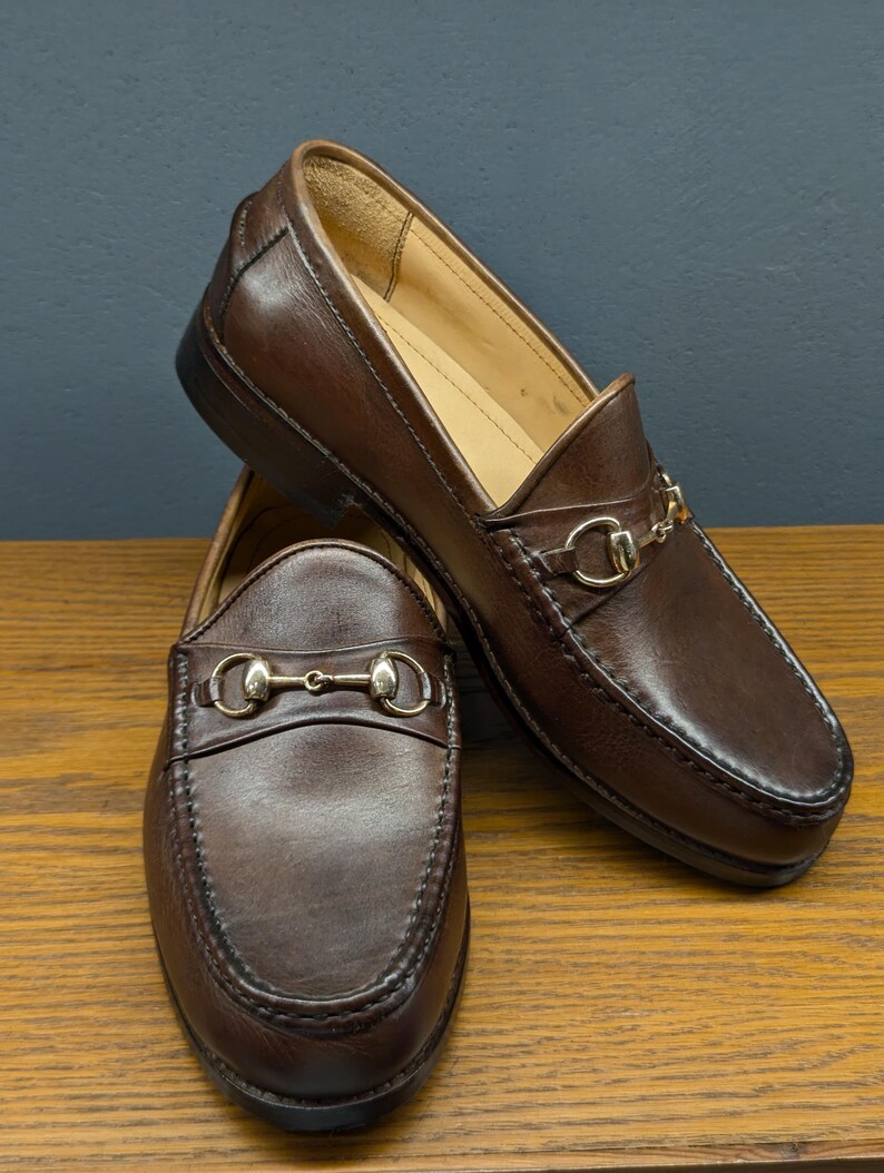 May include: A pair of brown leather loafers with gold-colored metal accents. The shoes are displayed on a wooden surface, showcasing their classic design and stitching details. The loafers feature a slip-on style with a decorative strap.