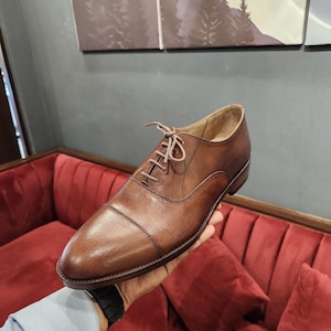 Peut inclure: Chaussure Oxford en cuir marron avec bout rapporté et fermeture à lacets. La chaussure a une finition polie et un design classique, adaptée aux tenues formelles ou professionnelles. La chaussure est tenue par une main.