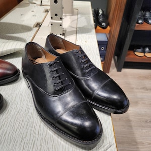 May include: A pair of black leather Oxford shoes with a cap toe. The shoes have a polished finish and are laced with black laces. They are displayed on a light-coloured surface, highlighting their sleek and classic design. The shoes are a classic style.